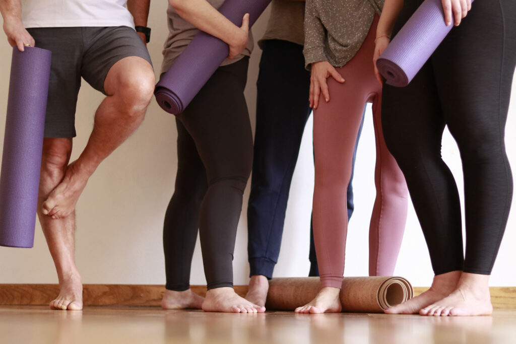 Man sieht die Beine von fünf Personen. Drei davon halten Yoga Matten in der Hand.